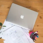 Top view of a laptop, papers, and pens on a wooden table in an office setting.